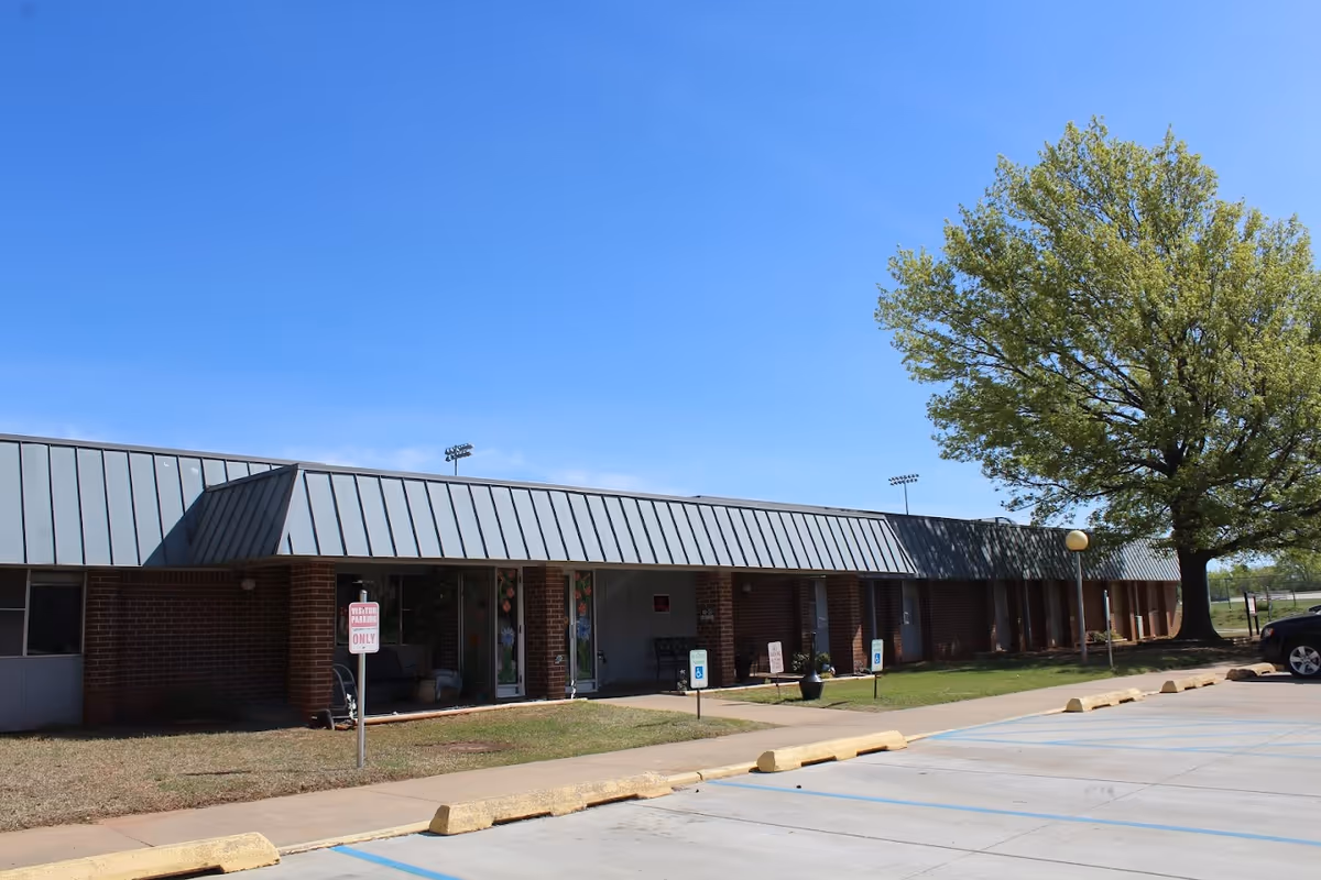 Exterior view of Shawnee Care Center, a single-story brick building with a metal roof. There is a large tree on the right side and a parking lot with designated handicapped parking spaces in front. The sky is clear and blue.