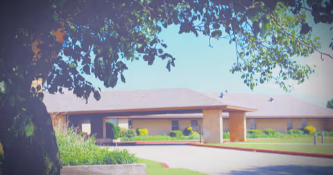 Exterior view of a single-story nursing center building with a covered entrance, surrounded by greenery and trees under a clear sky.