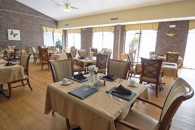 A bright dining room with multiple tables covered in beige tablecloths, each set with black napkins, white cups, and silverware. The room has large windows with yellow valances letting in natural light, brick walls, and wooden flooring. Chairs with striped upholstery surround the tables, and there are decorative plants and wall art visible.