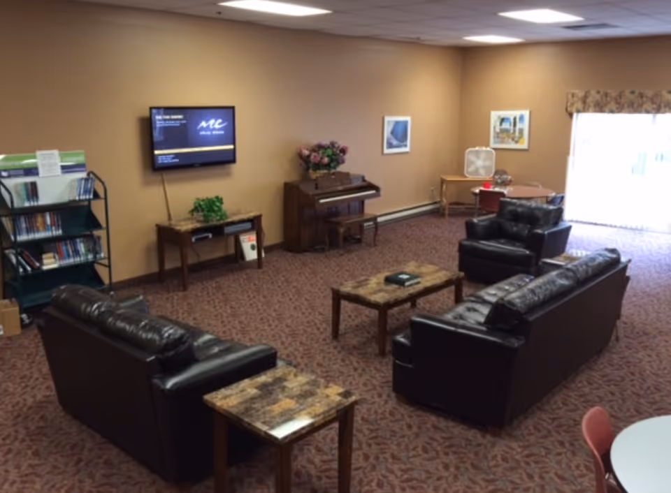 A senior living facility common area with two black leather sofas, a matching armchair, and two marble-top coffee tables arranged on a patterned carpet. A flat-screen TV is mounted on a beige wall above a small wooden table with a plant. There is a piano with a floral arrangement on top, a bookshelf with books, and a small table with chairs and a fan near a sliding glass door letting in natural light.