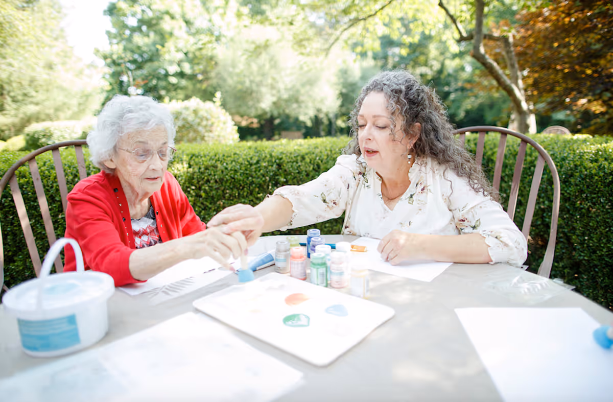 An elderly woman and a middle-aged woman sitting outdoors at a table engaged in a painting activity. The elderly woman is wearing a red cardigan and glasses, while the middle-aged woman with curly hair is helping her with the paintbrush. Various paint bottles and a palette with paint are on the table, with greenery and trees in the background.