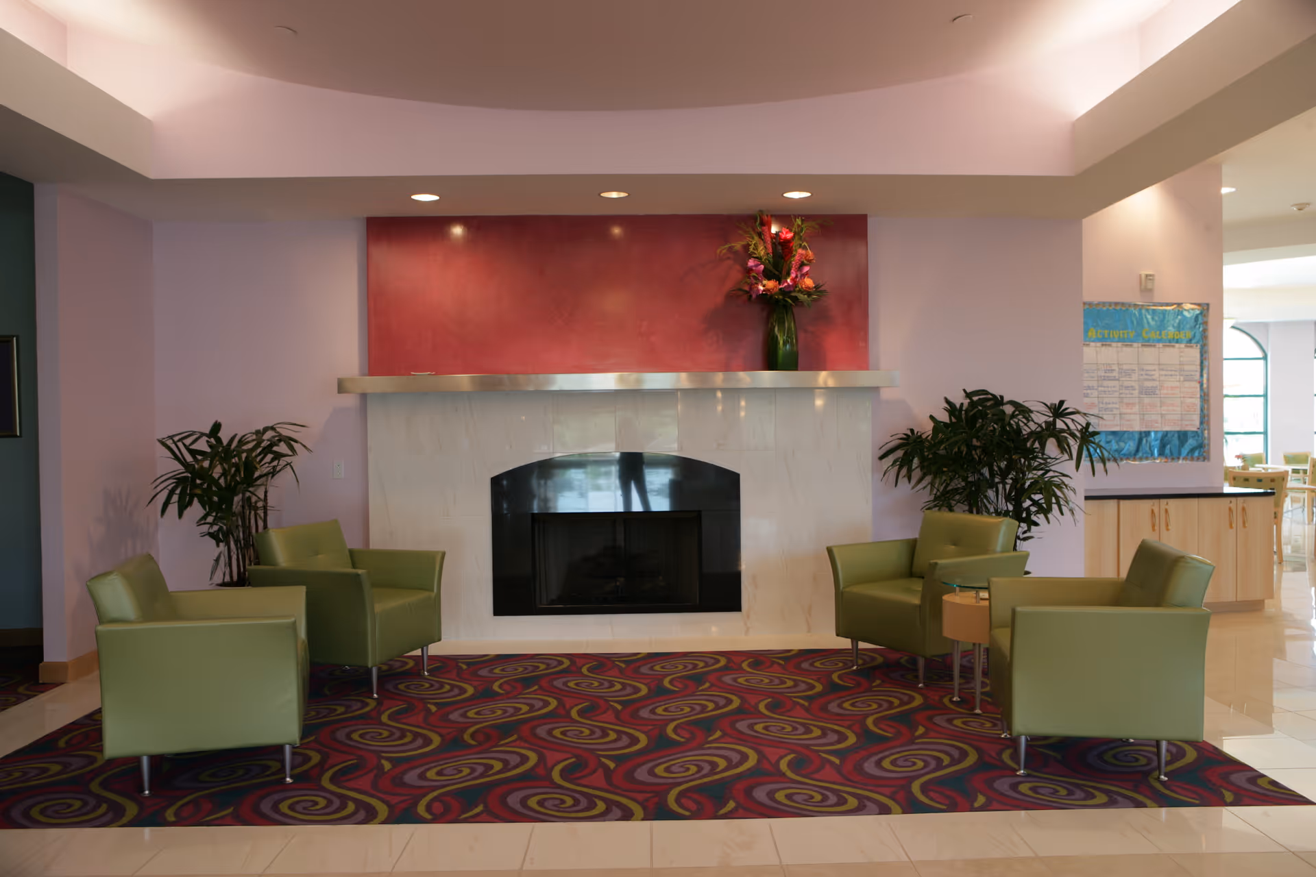 Lobby seating area with four green chairs arranged around a fireplace on a colorful patterned rug, flanked by plants and a vase of flowers on the mantel.