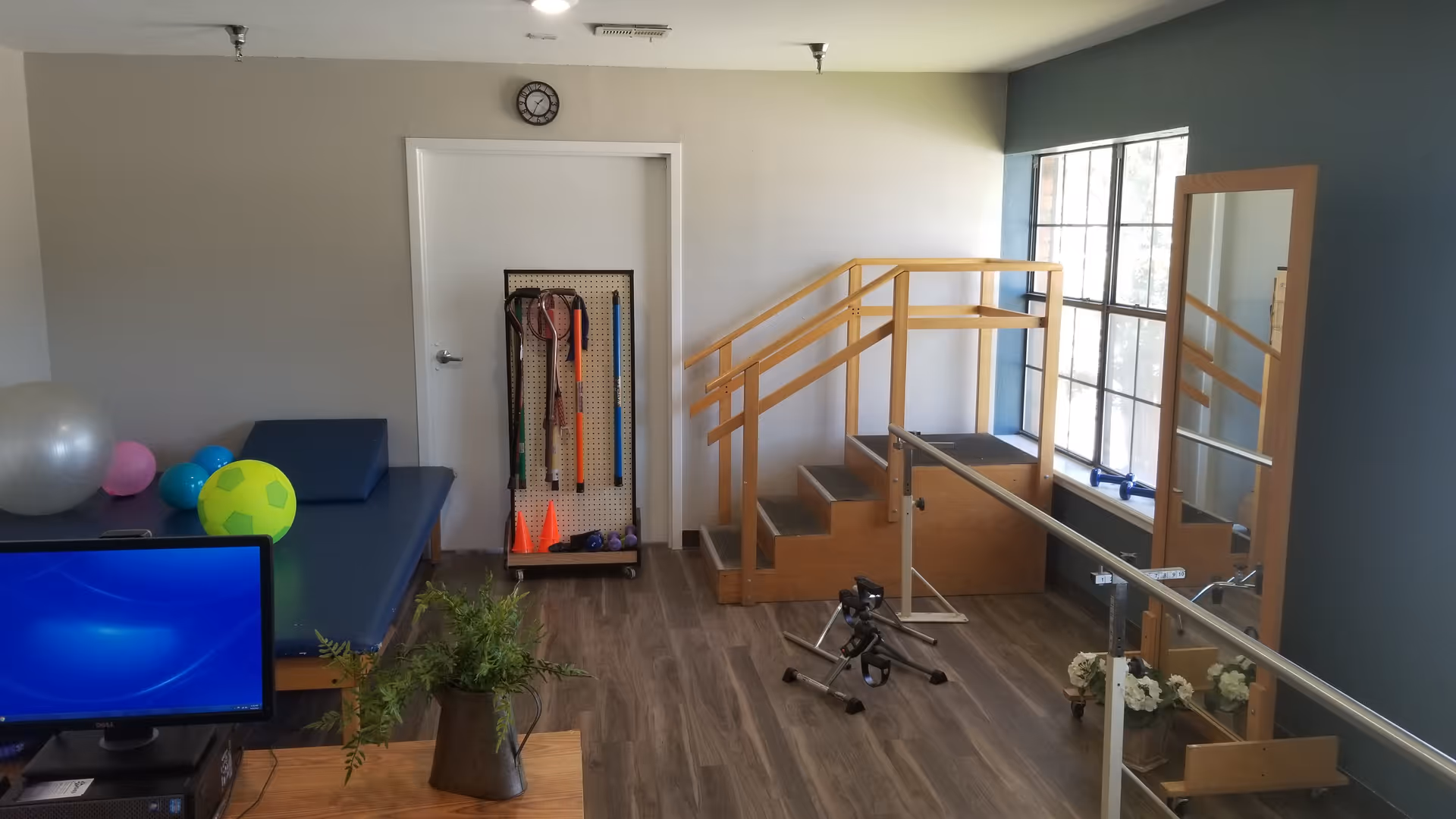 A physical therapy room with a blue therapy table holding various exercise balls, a set of wooden stairs with handrails for rehabilitation, a stationary pedal exerciser on the floor, a large mirror on the right wall, and a pegboard with exercise equipment such as cones and poles against the back wall. A computer monitor and a plant are visible in the foreground.