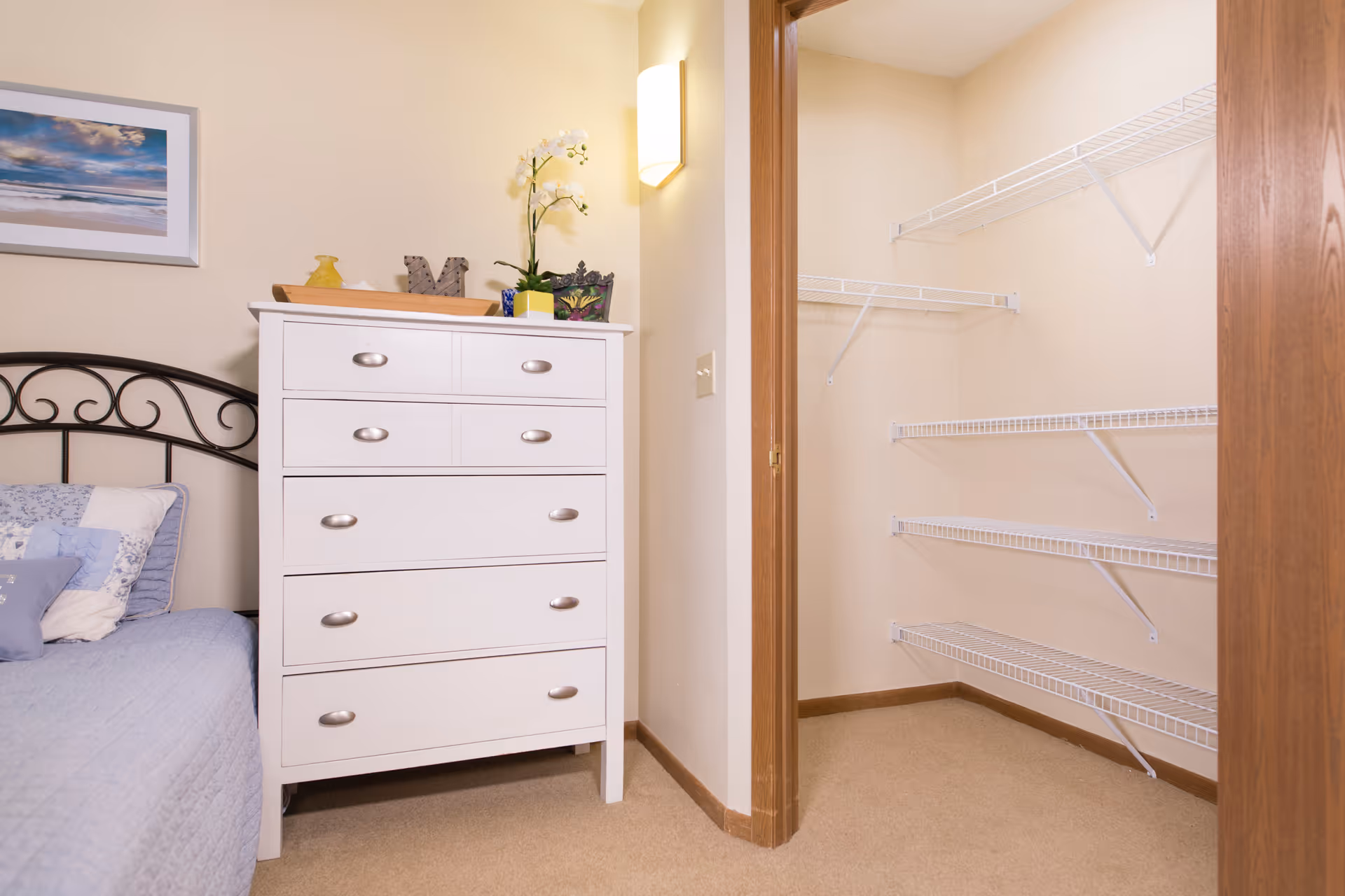 A bedroom corner with a white chest of drawers next to an open closet with wire shelving and part of a bed.