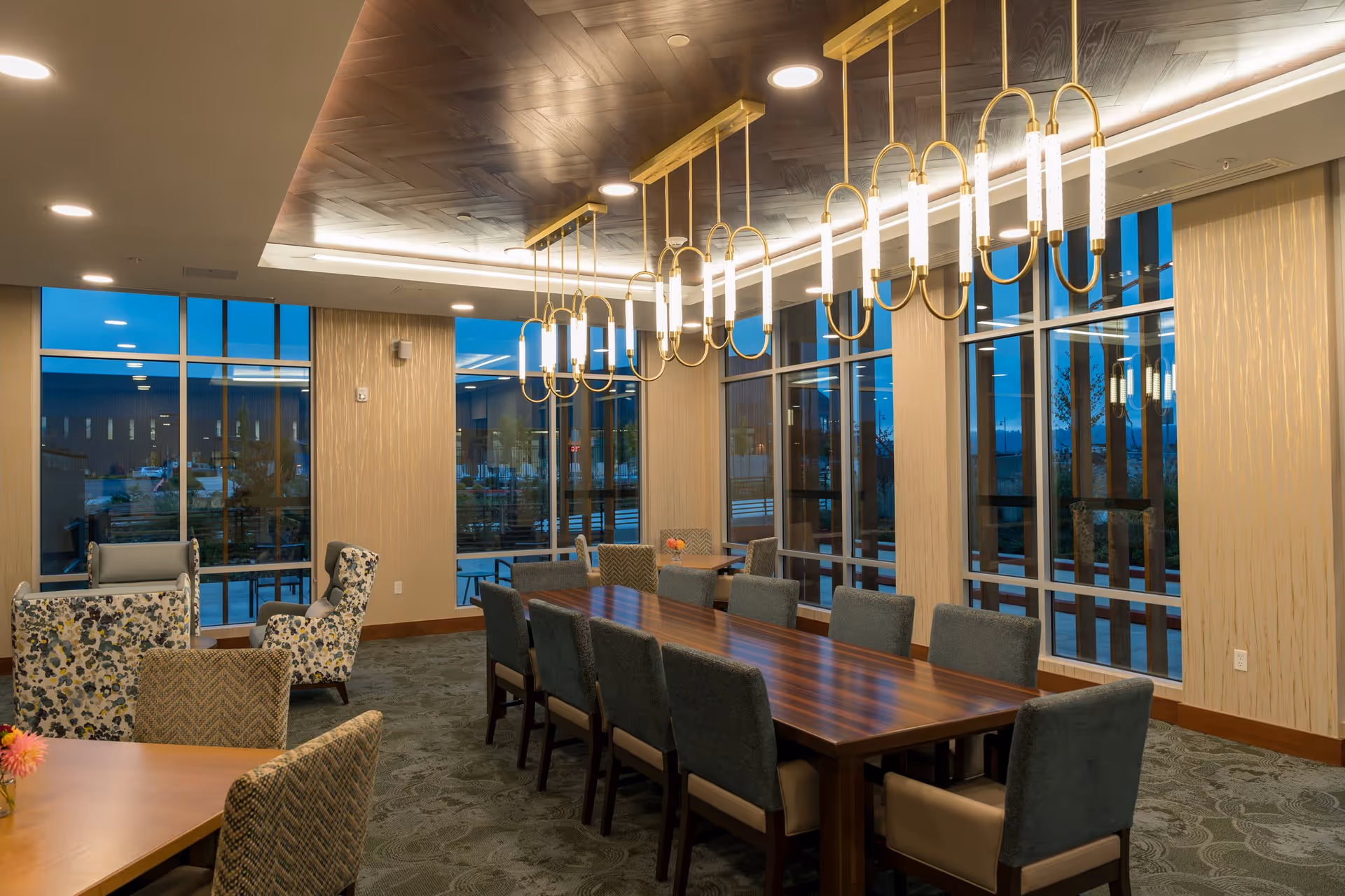 A spacious dining room with a long wooden table surrounded by cushioned chairs. The room features large floor-to-ceiling windows, modern pendant lighting fixtures hanging from a wooden ceiling, and additional seating areas with patterned armchairs. The setting appears elegant and well-lit during evening hours.