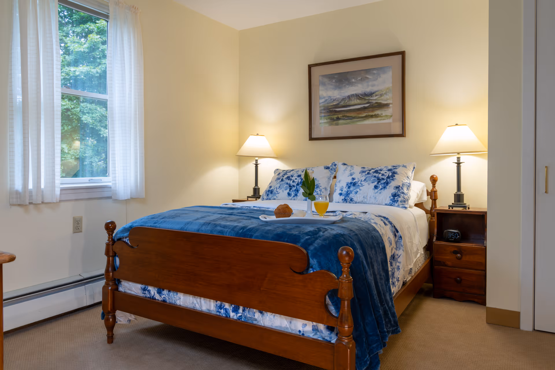 A cozy bedroom with a wooden bed frame, blue and white floral bedding, and a blue blanket. There are two bedside tables with lamps on each side of the bed. A framed landscape painting hangs on the wall above the bed. A window with white curtains allows natural light into the room.