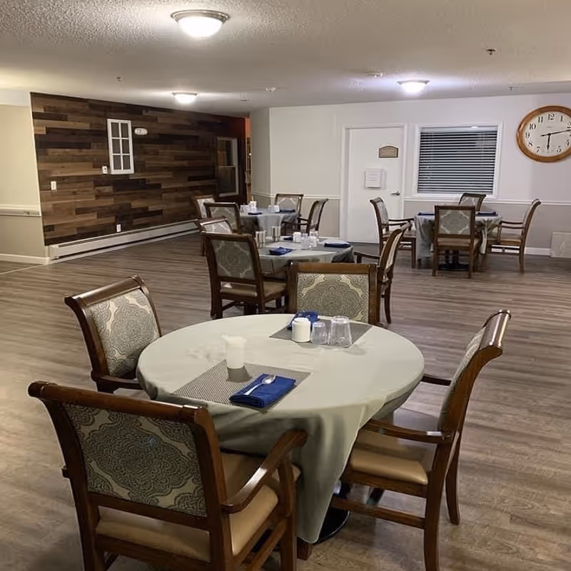 Communal dining room with round tables covered in tablecloths and wooden chairs in a senior living facility.