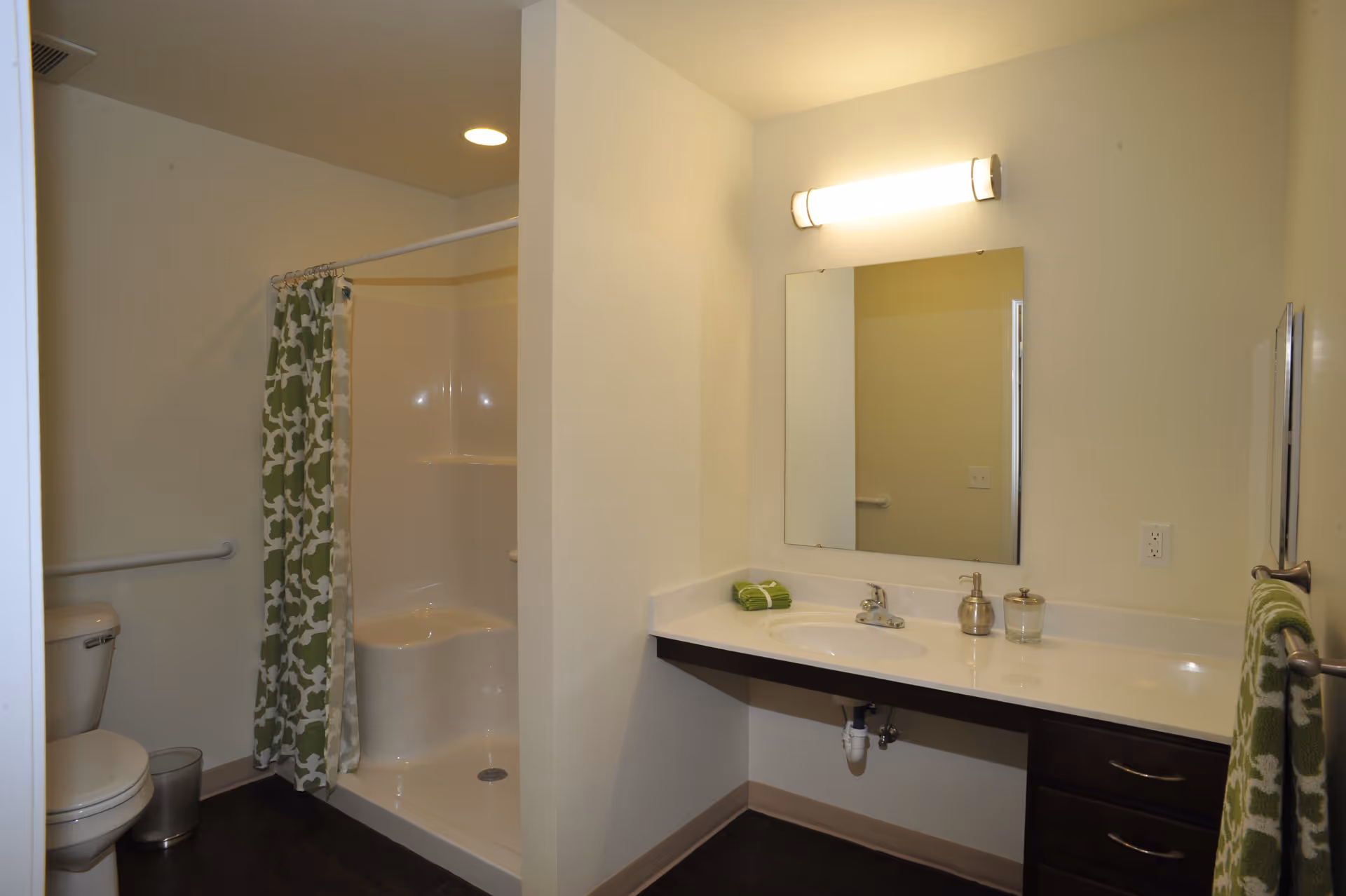 A bathroom with a white toilet, a shower with a green and white patterned curtain, a large mirror above a white countertop with a sink, and a green towel hanging on a rack. The walls are light-colored and the floor is dark.