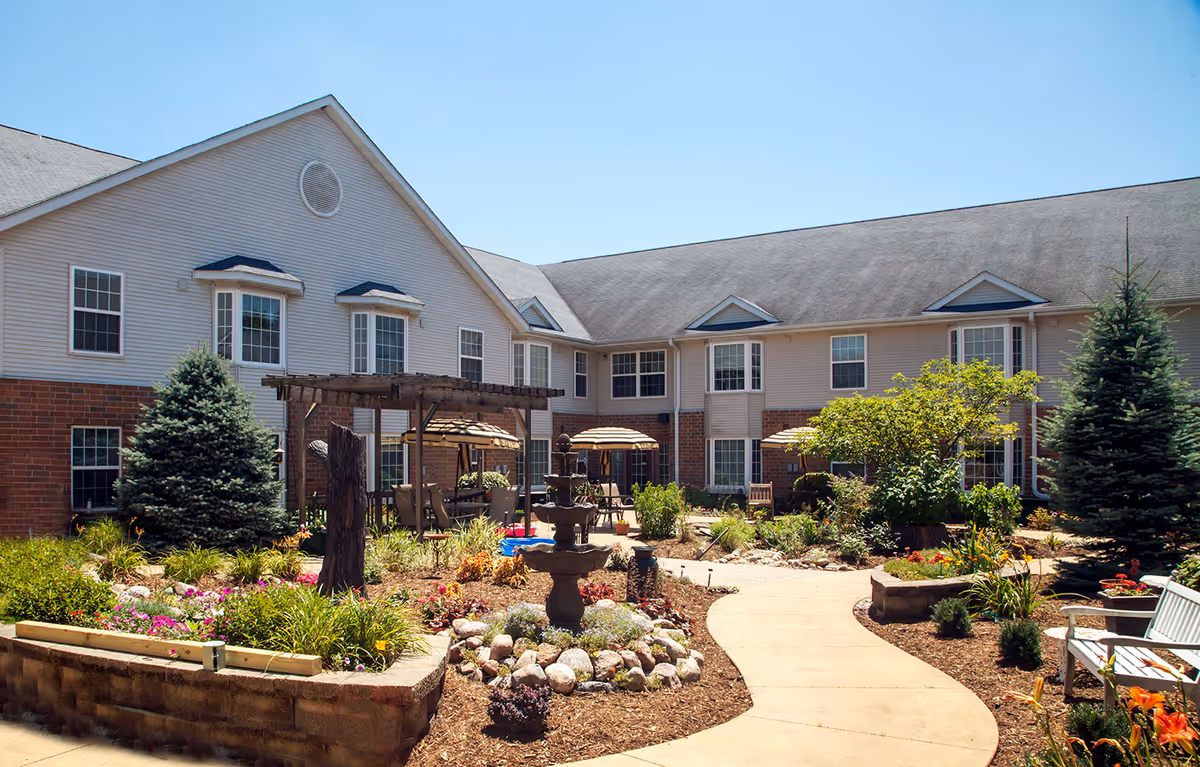 Outdoor courtyard area of Pennsylvania Place senior living facility featuring a paved walkway, garden beds with flowers and shrubs, a central water fountain, wooden pergola with seating, patio tables with umbrellas, and a two-story building with multiple windows in the background under a clear blue sky.