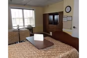 A nursing home bedroom with two beds, a window, a chair, and a wall-mounted TV cabinet.