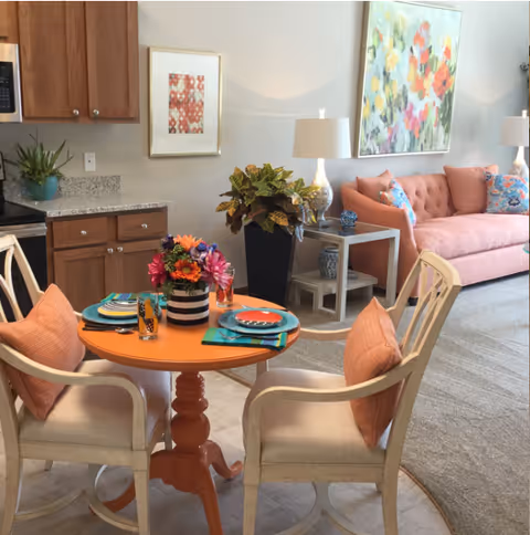 A cozy interior living space featuring a small round orange dining table set with colorful plates, glasses, and a striped vase with flowers. Two cushioned chairs with orange pillows surround the table. In the background, there is a peach-colored sofa with patterned pillows, a side table with a lamp and decorative items, a large colorful floral painting on the wall, and wooden kitchen cabinets with a granite countertop.