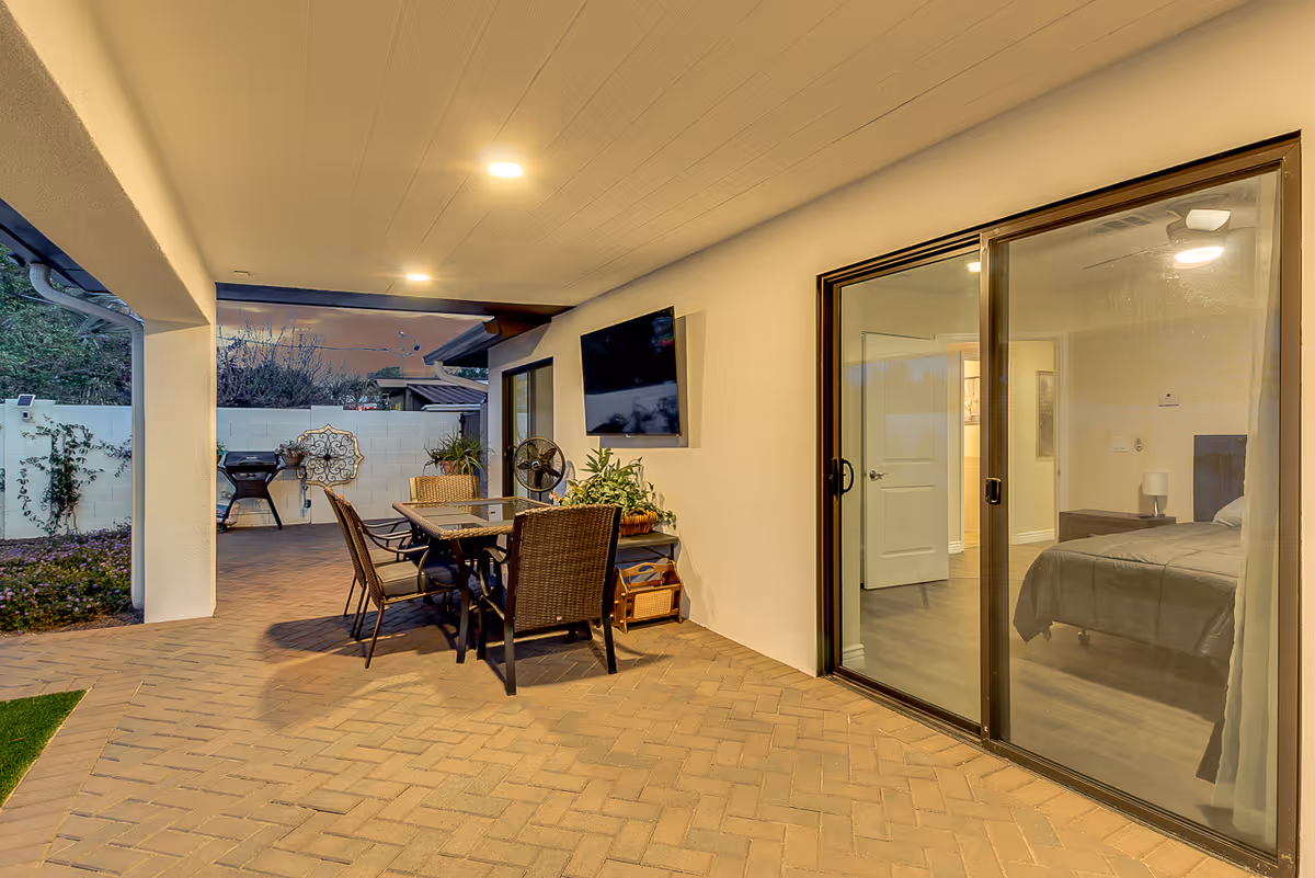 Covered outdoor patio area with a dining table and chairs, a wall-mounted TV, potted plants, and a sliding glass door leading to a bedroom inside. The patio has brick flooring and ceiling lights, with a barbecue grill and decorative wall art visible in the background.
