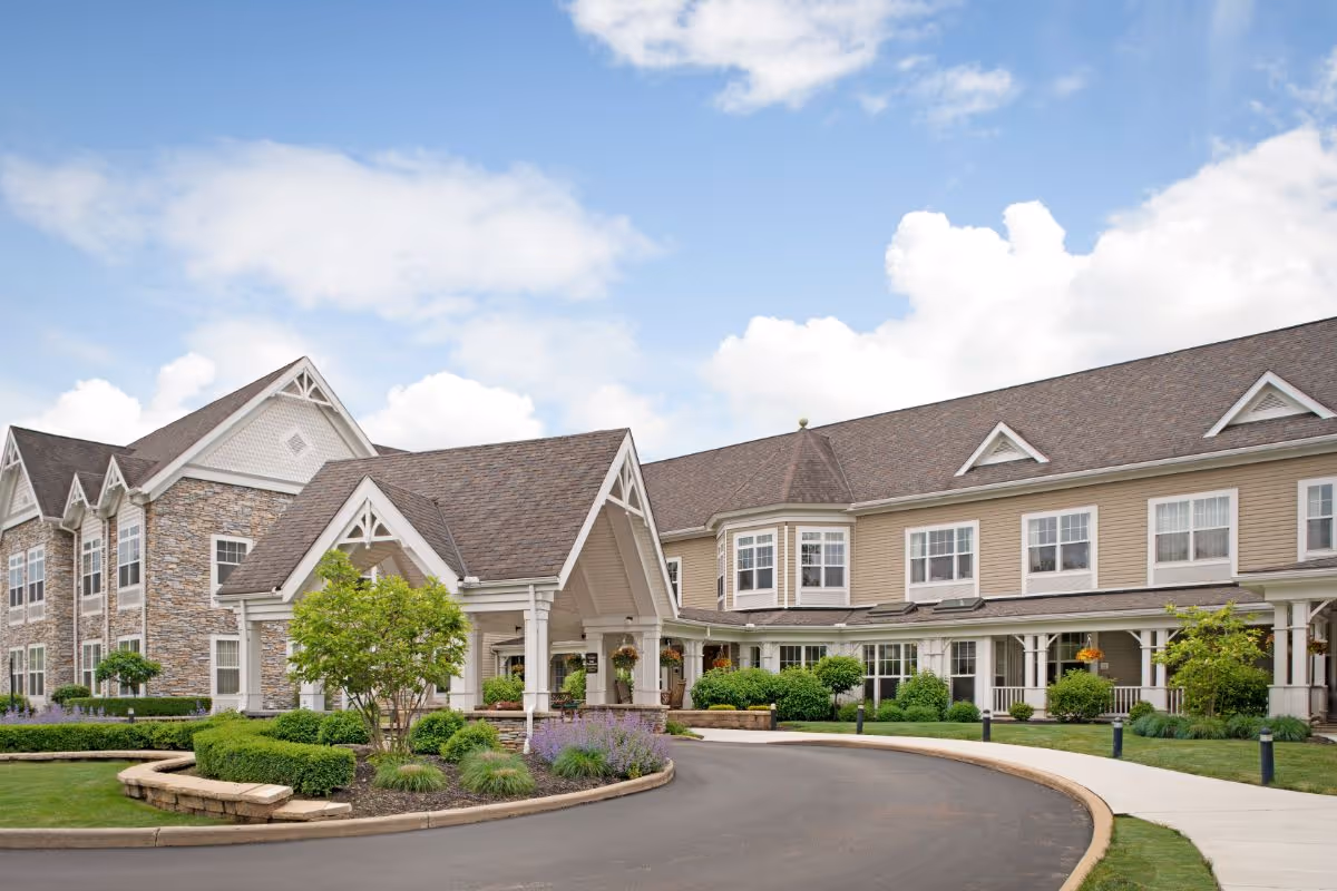 Front entrance of a two-story senior living building with a covered porte-cochere, landscaped circular driveway and blue sky.