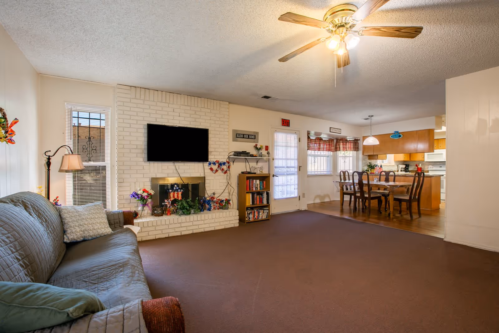 A cozy living room with a brown couch covered by a quilted cover, a floor lamp, and a white brick fireplace with a mounted flat-screen TV above it. The fireplace mantel is decorated with flowers and patriotic-themed decorations. To the right, there is a dining area with a wooden dining table and six chairs, a hanging light fixture, and windows with red checkered curtains. The room has a ceiling fan with lights and a brown carpeted floor, transitioning to wood flooring in the dining area.