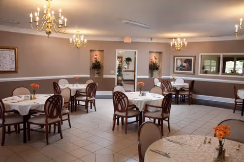 Dining room with round tables and chairs, chandeliers, and small floral centerpieces.