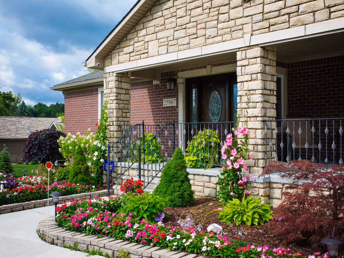 Front entrance of a brick and stone building with a covered porch, decorative railing, and a well-maintained garden featuring various colorful flowers and shrubs.
