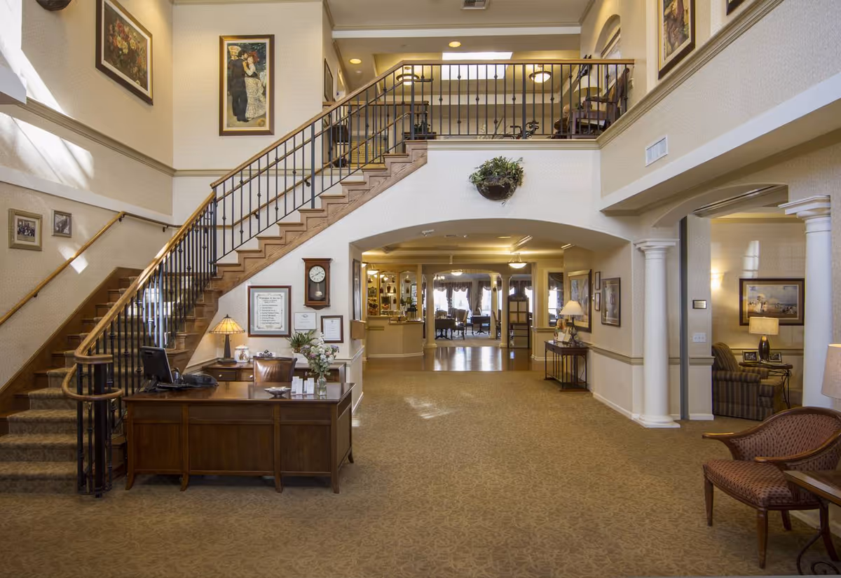 Spacious and warmly lit interior of a senior living facility lobby with a wooden reception desk, a staircase with a black metal railing leading to an upper floor, framed artwork on the walls, and a seating area visible through an archway. The decor includes lamps, plants, and classic furniture, creating a welcoming atmosphere.