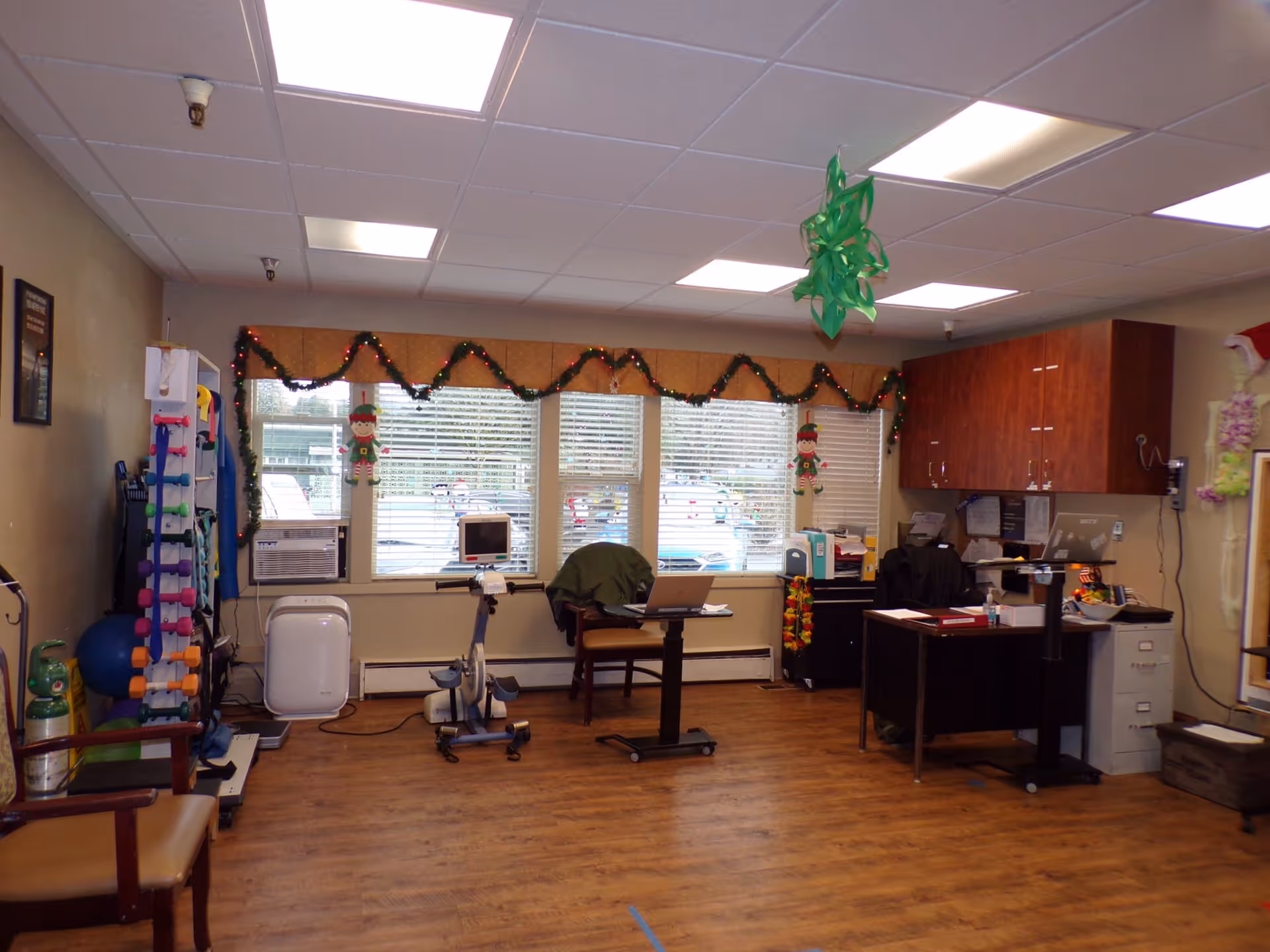 A room with wooden flooring and beige walls, featuring exercise equipment including a stationary bike and colorful dumbbells on a rack. There are three large windows with blinds, decorated with garlands and hanging elf ornaments. A desk with office supplies and a chair is positioned near the windows, and wooden cabinets are mounted on the wall. The ceiling has fluorescent lights and a green hanging decoration.