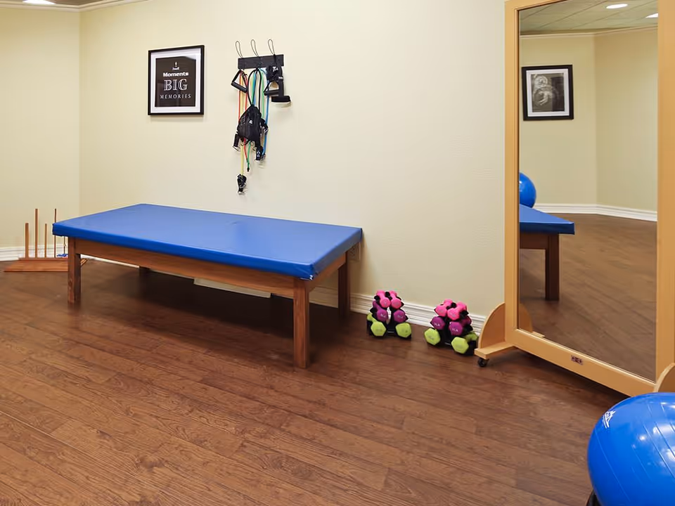 A small exercise room with a blue padded bench, resistance bands hanging on the wall, two sets of pink and green dumbbells on the floor, a large standing mirror, and a blue exercise ball partially visible. The room has wooden flooring and light-colored walls with framed pictures.