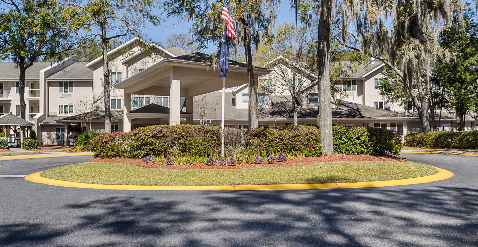 Front exterior of a senior living facility showing a covered entrance/porte-cochère, flagpole, circular drive and landscaped island with trees.