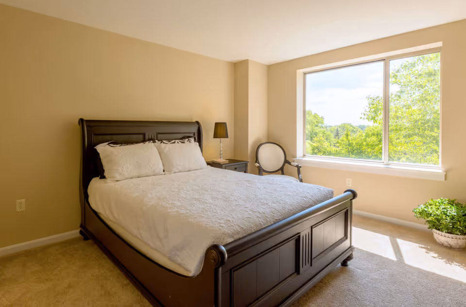Sunlit bedroom with a dark wood bed dressed in white linens, a nightstand and lamp, a chair, and a large window overlooking trees.