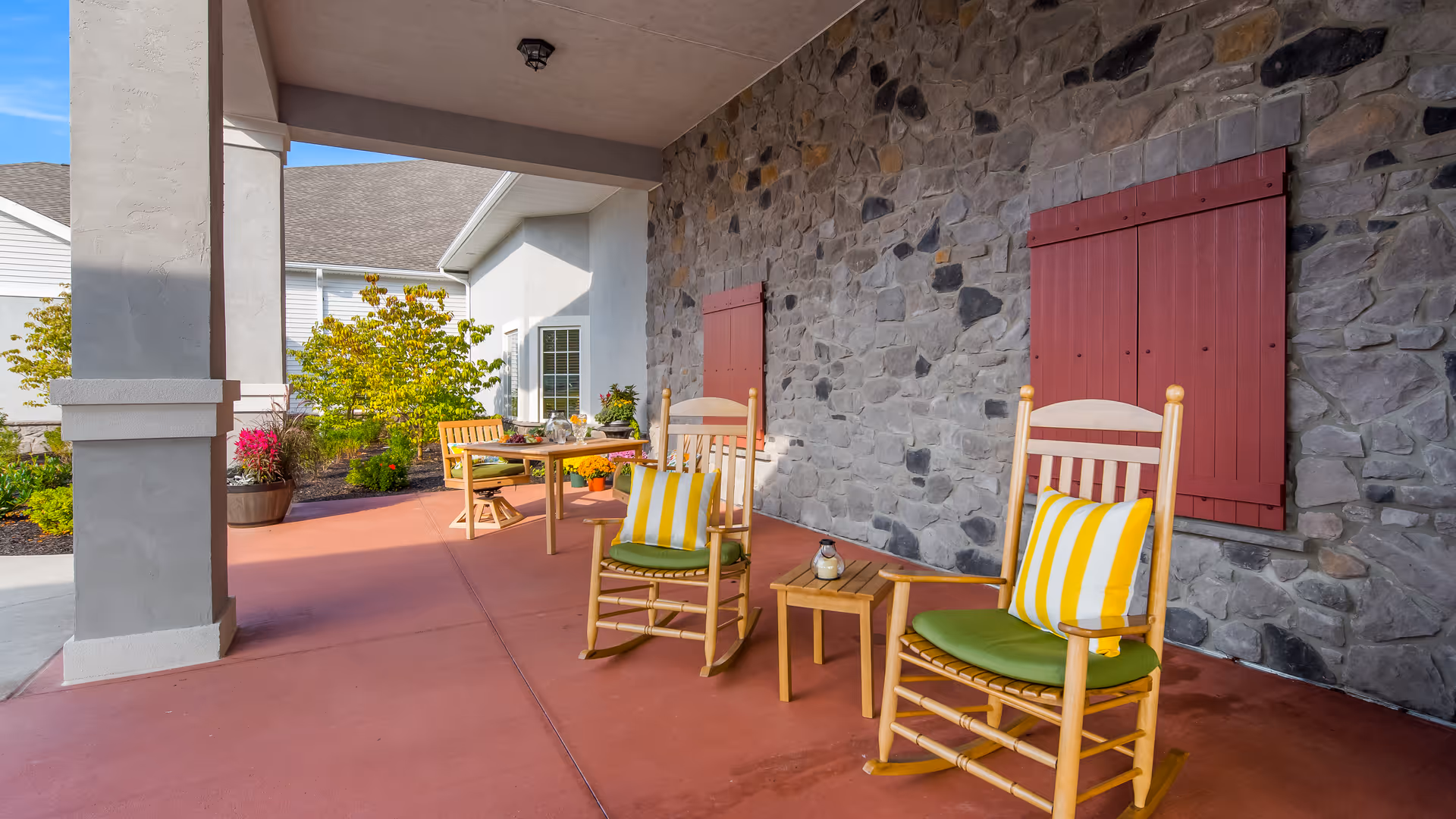 Outdoor covered patio area with two wooden rocking chairs featuring green cushions and yellow striped pillows, a small wooden side table between them, and a wooden dining table with chairs in the background. The patio has a stone wall with two red wooden shutters and potted plants nearby.
