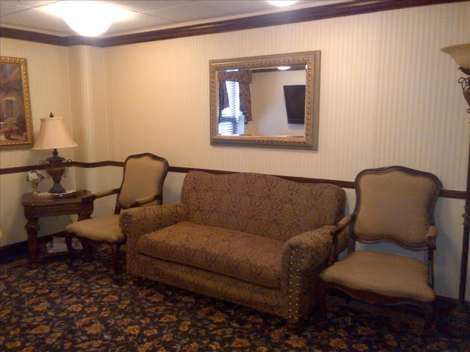 A cozy living room area with a patterned brown sofa flanked by two upholstered chairs. There is a wooden side table with a decorative lamp and a small flower arrangement on the left. A large ornate mirror hangs on the wall above the sofa, reflecting a window with blinds and a television. The room has beige striped wallpaper, dark wood trim, and a patterned carpet.