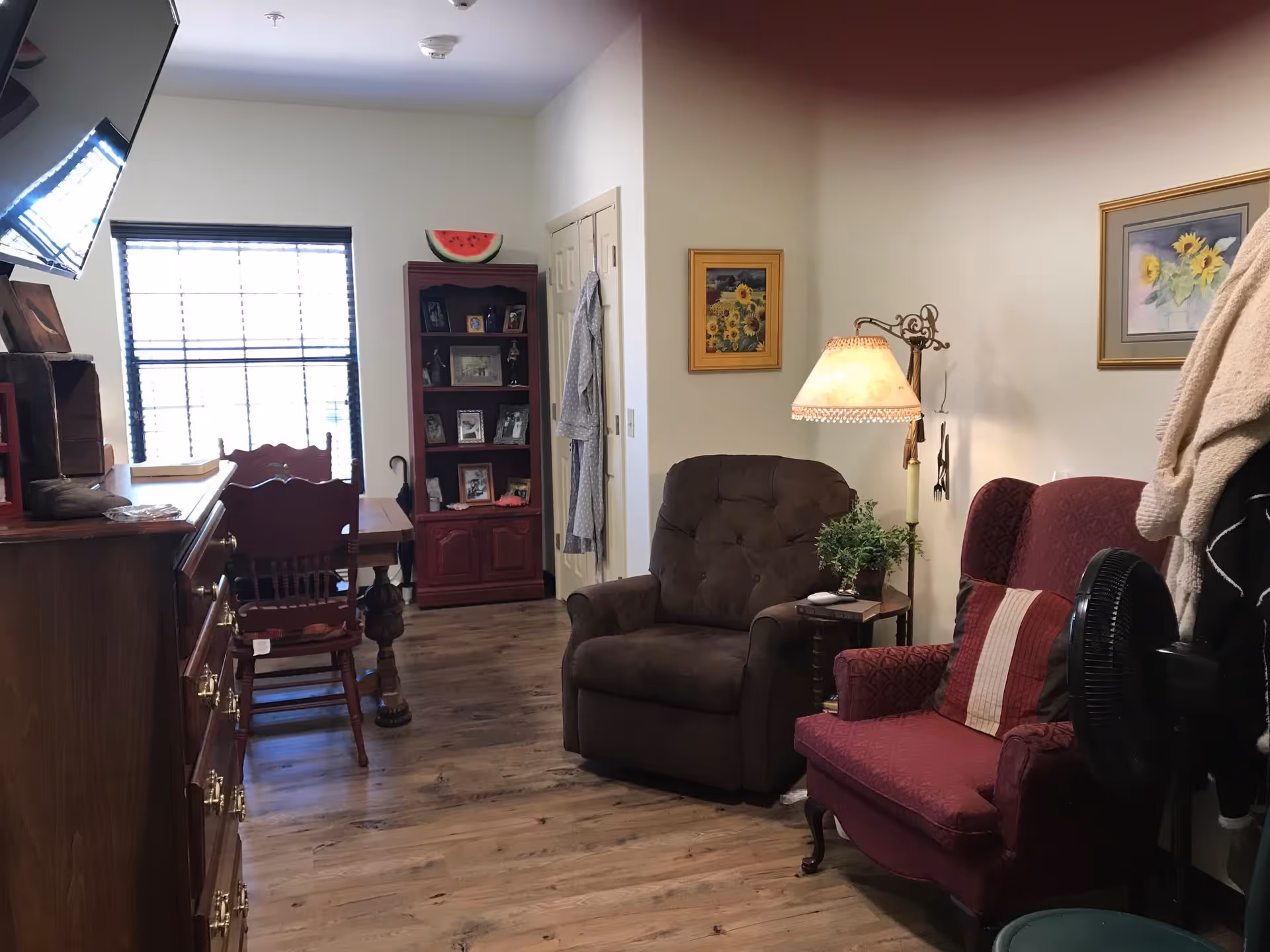 Cozy living area with a brown recliner and a red armchair with a striped pillow. A floor lamp with a decorative shade stands between the chairs. On the wall are two framed sunflower paintings. To the left, there is a wooden dining table with chairs near a window with blinds. A wooden cabinet with framed photos and a watermelon decoration on top is against the back wall. The floor is wood, and the walls are light-colored.