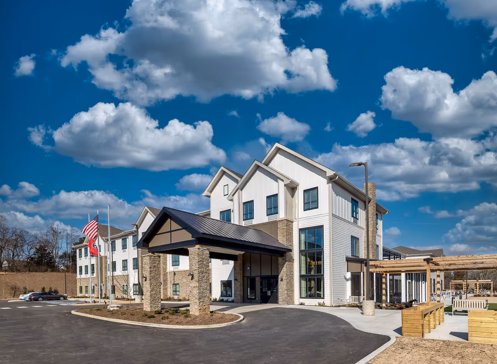 Front exterior of a three-story senior living building with a covered porte-cochere, flagpoles, and outdoor seating under a blue sky.