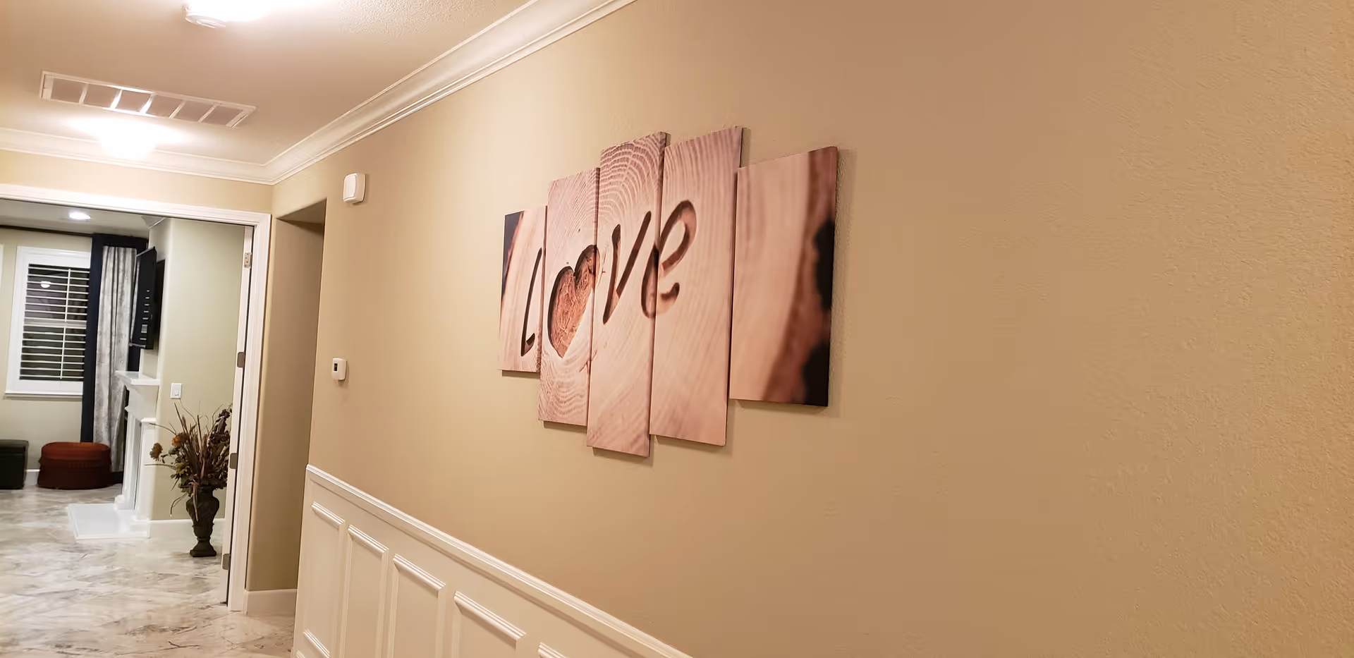 A hallway with beige walls and white wainscoting, featuring a multi-panel wall art piece spelling the word 'LOVE' with a heart shape in place of the letter O. At the end of the hallway, there is an open doorway leading to a living room area with a fireplace, a mounted TV, a window with curtains, and some furniture including a red ottoman and a green ottoman.