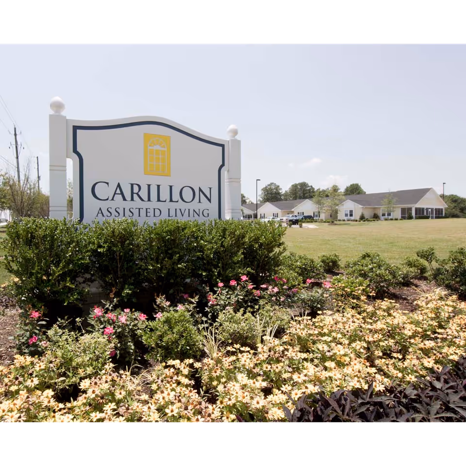 Outdoor view of Carillon Assisted Living of Clemmons sign surrounded by bushes and flowers, with the assisted living buildings visible in the background under a clear sky.