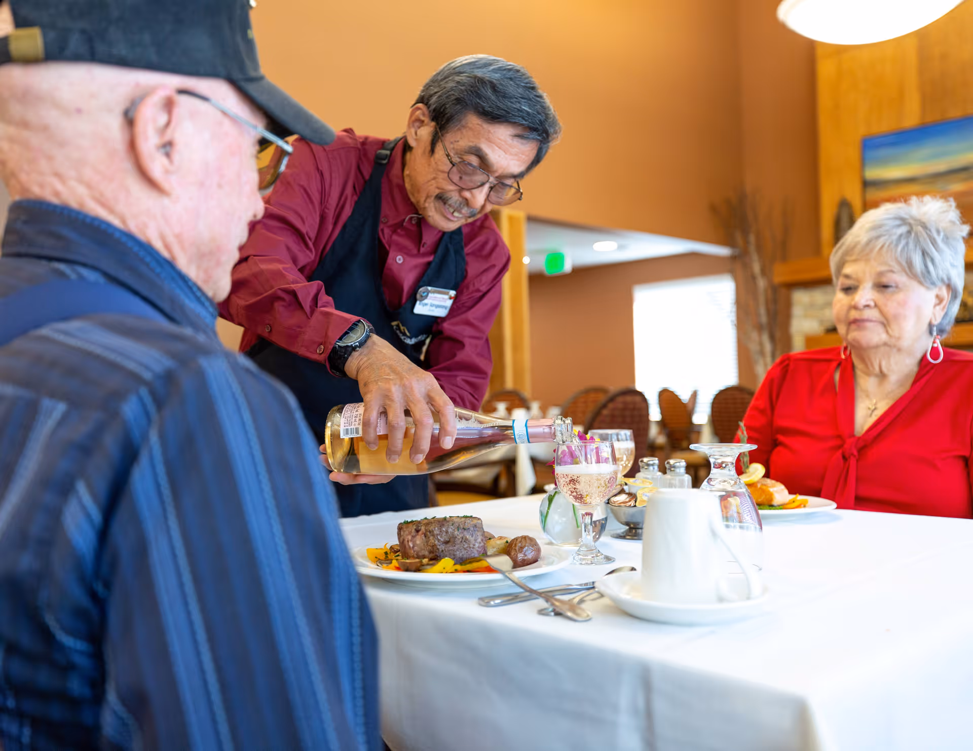 A server wearing a red shirt and black apron is pouring a drink into a glass at a dining table where an elderly man and woman are seated. The table is set with plates of food, utensils, and glasses in a warmly lit dining room.