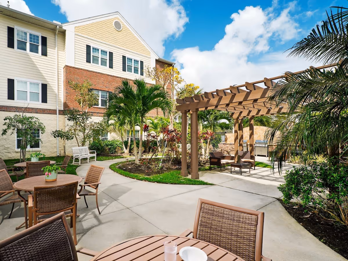 Outdoor patio area at a senior living facility with round tables and chairs, a wooden pergola with seating underneath, surrounded by palm trees and other greenery, with a multi-story building in the background under a partly cloudy blue sky.