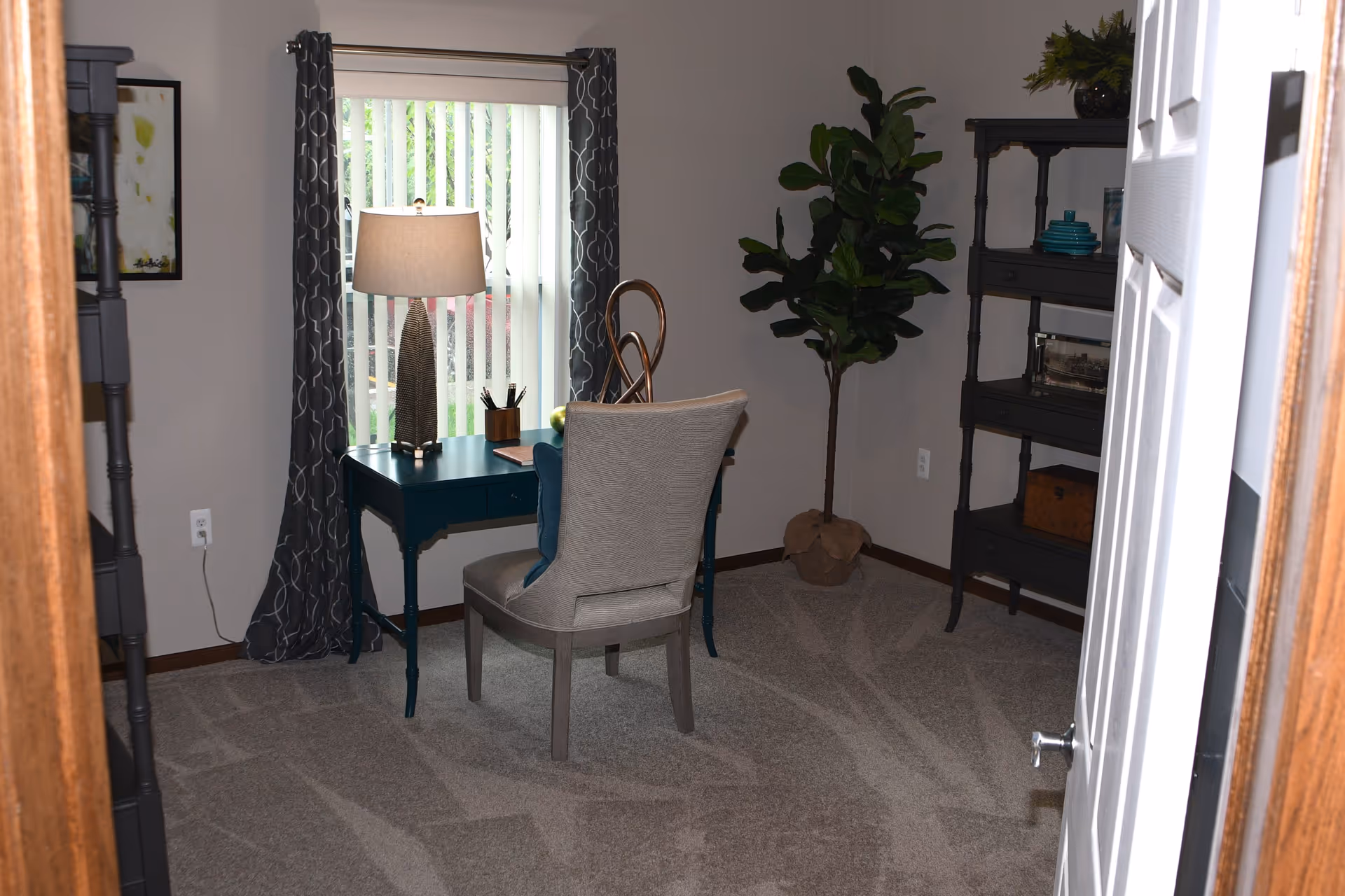 A small room with a gray carpeted floor featuring a dark teal desk with a lamp, a chair with a cushion, a tall potted plant in the corner, and a dark shelving unit with decorative items. The window has vertical blinds and patterned curtains.