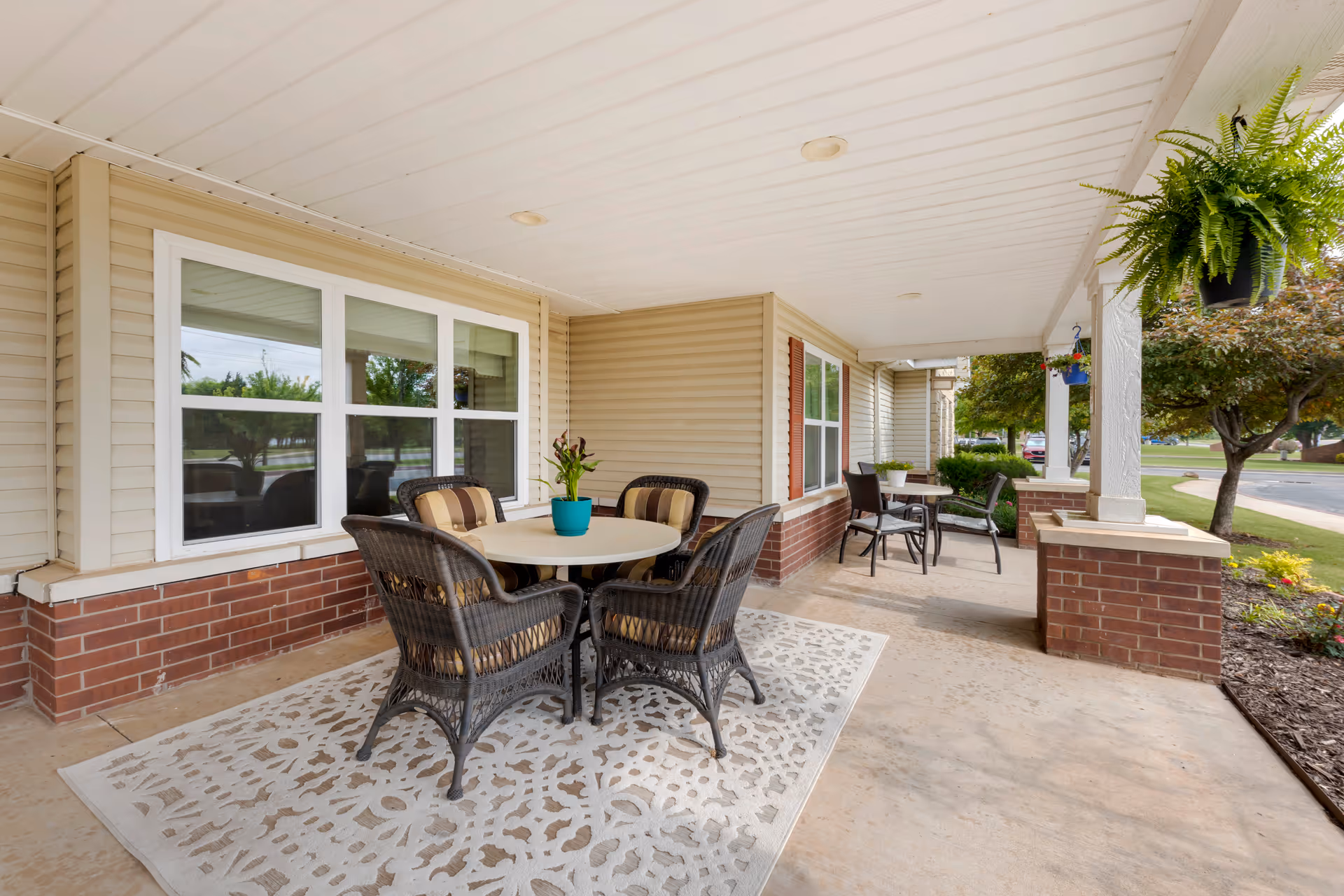 Covered outdoor patio area with two round tables, each surrounded by four chairs. One table has a potted plant in a blue pot, and the other has a small white pot. The patio has a white ceiling with recessed lighting, beige siding walls with brick accents, and hanging plants. Trees and a road are visible in the background.