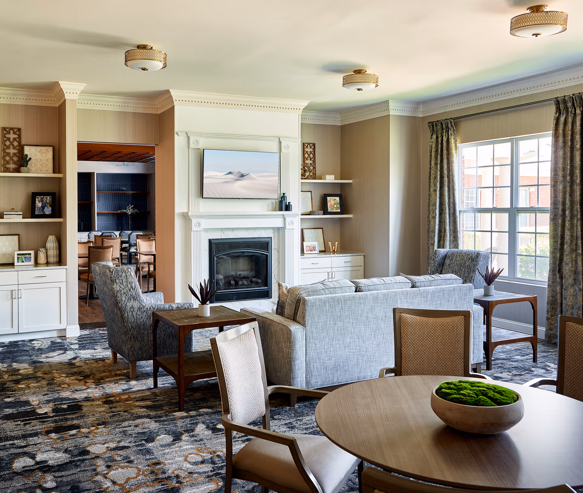 A bright and spacious living room area in a senior living facility with a large window letting in natural light. The room features a gray sofa, patterned armchairs, a round wooden table with chairs, and a fireplace with a mounted TV above it. Built-in shelves with decorative items and framed pictures flank the fireplace. The carpet has a patterned design, and curtains frame the window.