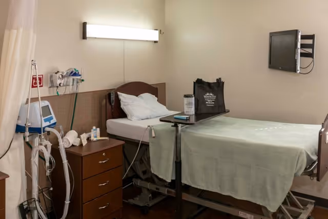 A hospital-style patient room with a single bed covered with a light green blanket and a white pillow. Next to the bed is a small wooden nightstand with medical supplies and equipment. A medical monitor and other devices are mounted on the wall. A black bag and a water bottle are placed on an overbed table. A flat-screen TV is mounted on the wall opposite the bed. The room has beige walls and a curtain partially visible on the left side.