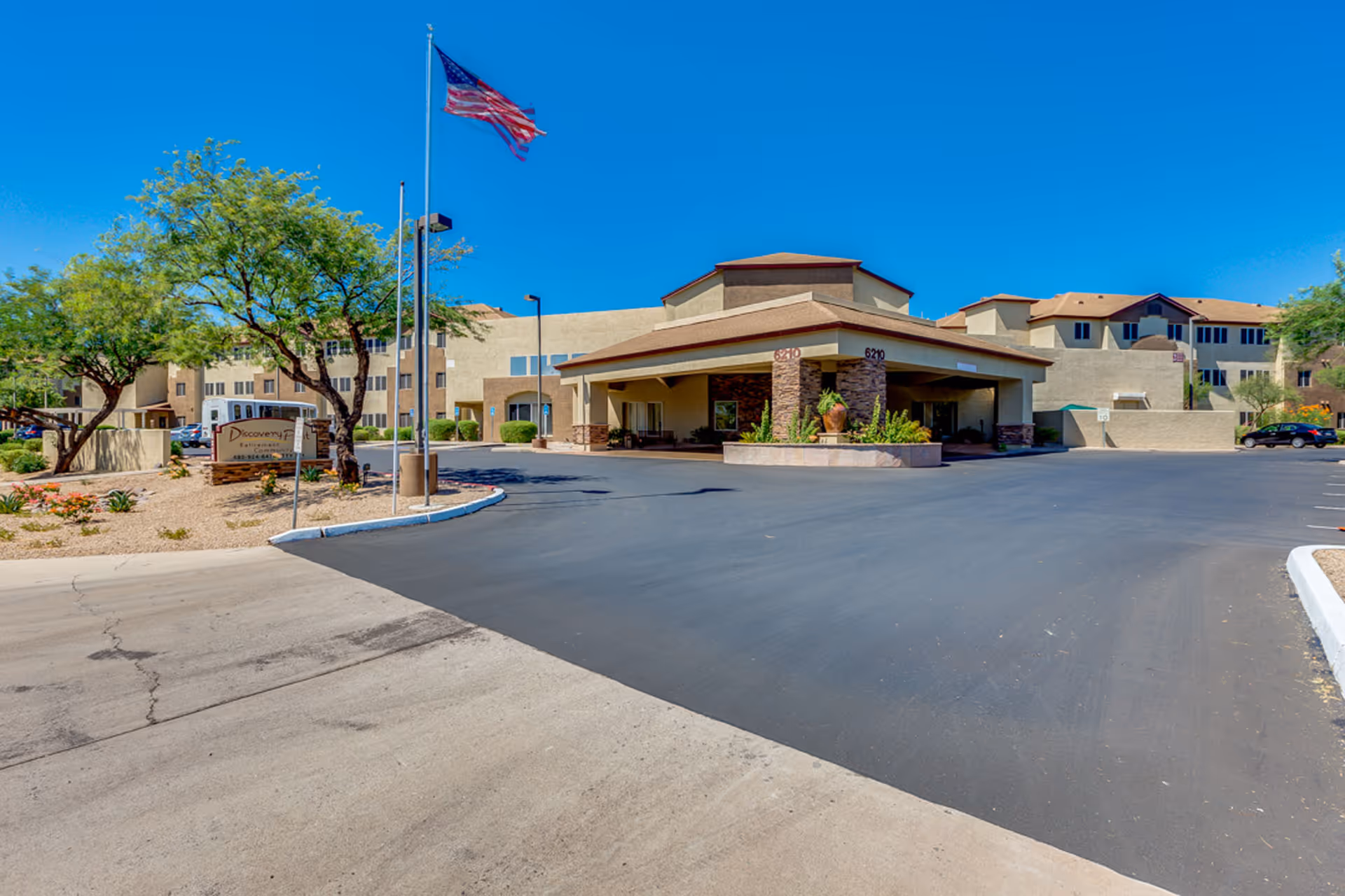 Exterior view of Discovery Point Retirement Community building with a driveway, landscaped area with trees and shrubs, an American flag on a flagpole, and clear blue sky.