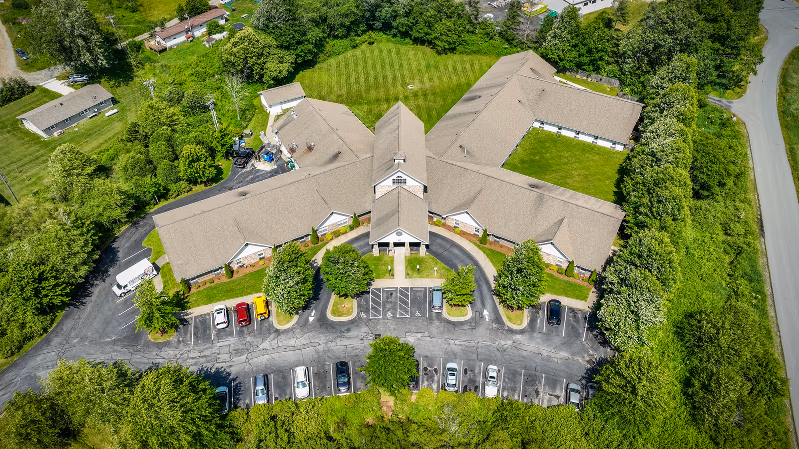 Aerial view of Deerfield Ridge Assisted Living facility showing a large, single-story building with a beige roof surrounded by green lawns, trees, and a parking lot with several cars parked. The building is shaped like a wide 'M' with a central entrance and driveways leading to the parking area.