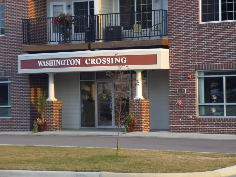 Entrance of a brick building with a sign above the door that reads 'WASHINGTON CROSSING'. There are two white columns supporting the sign, potted plants on either side of the entrance, and a small tree in front of the building.