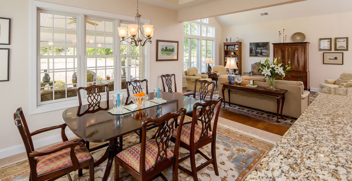 A bright and spacious interior featuring a dining area with a polished wooden table and six chairs with patterned cushions. The dining area is adjacent to a living room with beige sofas, armchairs, a wooden cabinet, and decorative lamps. Large windows allow natural light to fill the space, and a granite countertop is visible in the foreground.