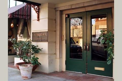 Entrance to the Evergreen Hotel with green double doors, a sign on the wall reading 'Evergreen Hotel', potted plants on the sidewalk, and a striped awning above a window.