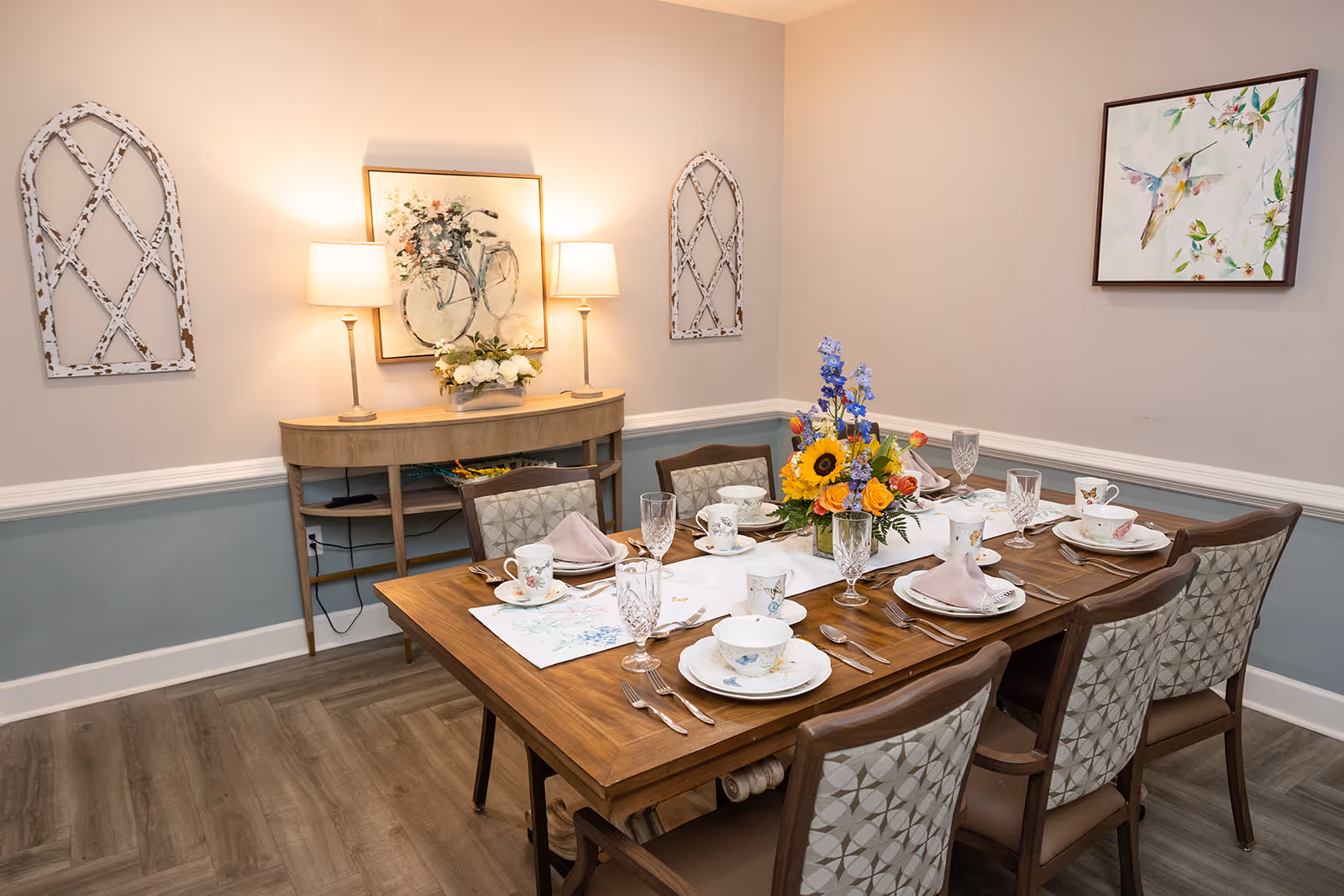 A dining room with a wooden table set for six people, featuring floral-patterned china, crystal glasses, and folded napkins. The table has a centerpiece with colorful flowers including sunflowers and delphiniums. The room has light beige walls with a blue wainscot, two table lamps on a wooden sideboard, and decorative wall art including a painting of a bicycle with flowers and a hummingbird artwork.