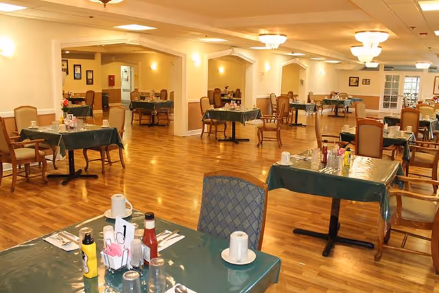 A spacious dining room with multiple tables covered in green tablecloths, each set with cups, condiments, and utensils. The room has wooden flooring, beige walls, and ceiling lights providing warm illumination. Chairs with cushioned seats surround the tables, and the area appears clean and well-organized.