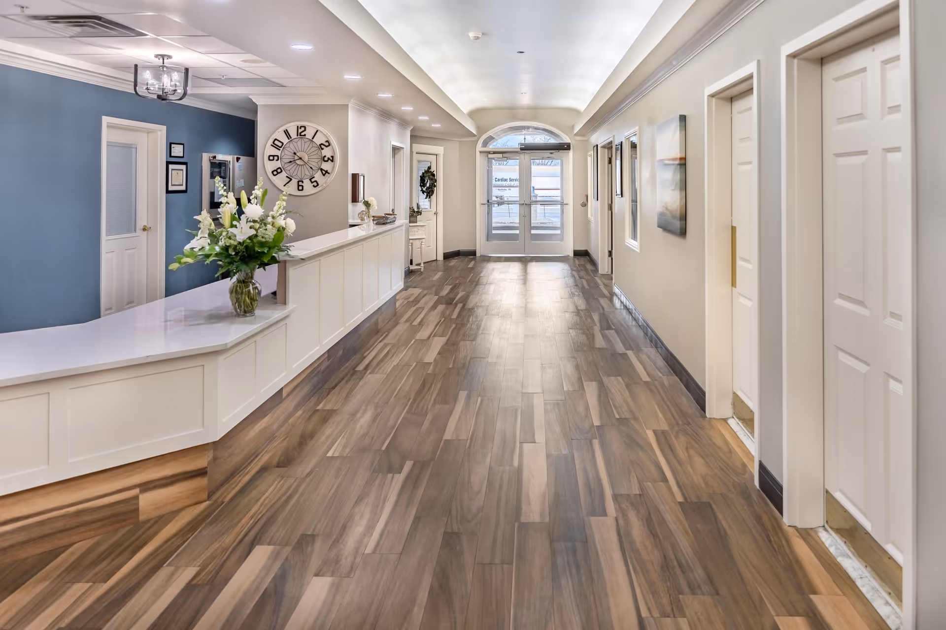 A bright and clean hallway in an assisted living facility with wood-patterned flooring, white walls, and multiple white doors along the right side. On the left side, there is a white reception desk with a vase of white flowers. A large clock is mounted on the wall behind the desk, and double glass doors are visible at the end of the hallway.