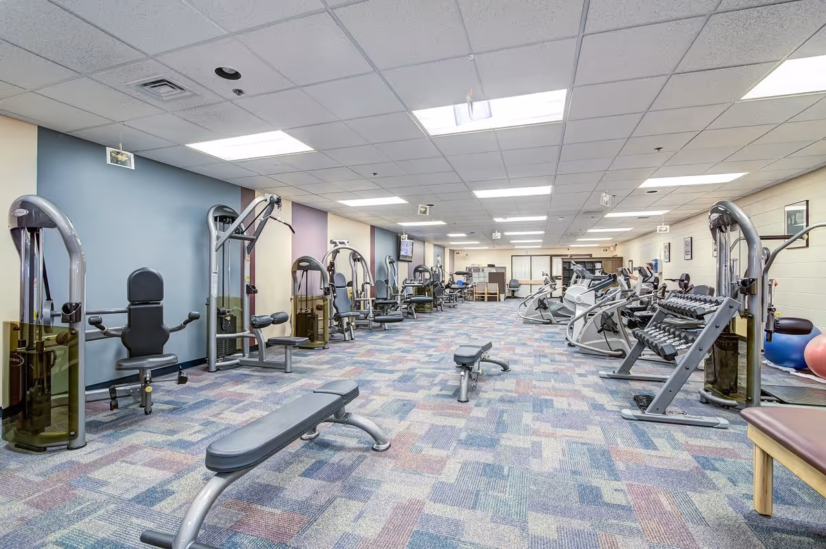 A spacious fitness room with various exercise equipment including weight machines, benches, dumbbells, and cardio machines. The room has a patterned carpet floor, light-colored walls with some sections painted in blue and purple, and a drop ceiling with fluorescent lighting.