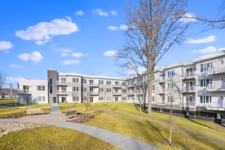 Courtyard view of a multi-story white senior living building with balconies, walkways, benches, and a large tree under a blue sky.