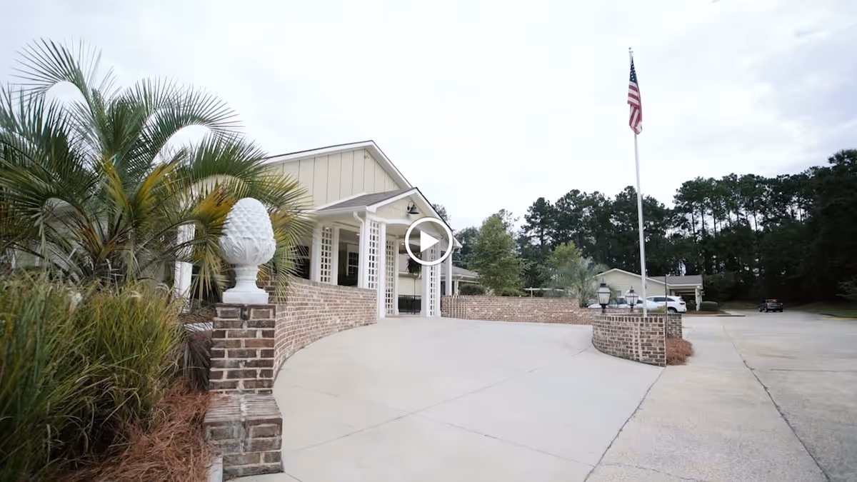 Entrance driveway to Fellowship Cordelia Manor Assisted Living Community featuring a curved brick wall with decorative white pineapple finials, palm trees and other greenery on the left, an American flag on a flagpole on the right, and a beige building with a covered porch in the background.