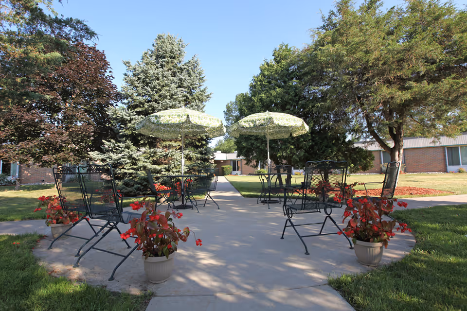 Outdoor seating area with metal chairs and tables under green patterned umbrellas, surrounded by potted red flowers and trees, with a building in the background under a clear blue sky.