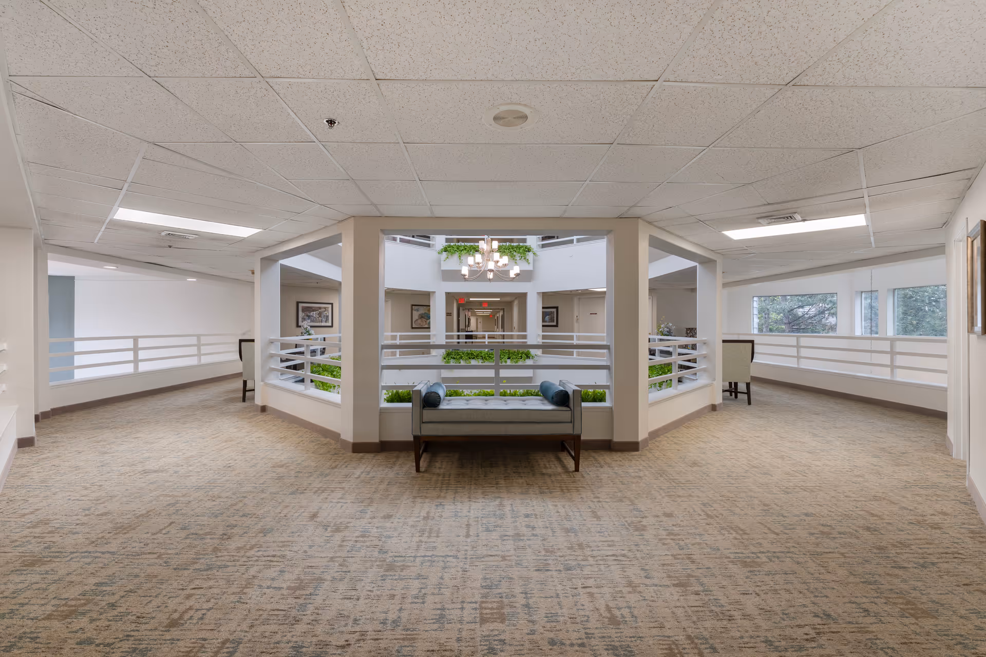 Bright open interior mezzanine atrium with a bench and railings overlooking a planted central courtyard and chandelier.